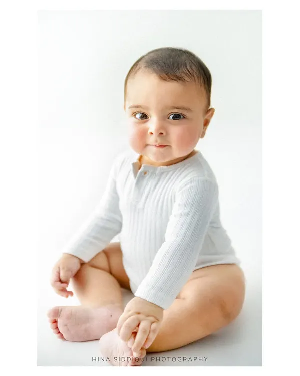 The sweetest six month smile. This portrait captures the adorable chubby cheeks and bright eyes of a baby at their half birthday milestone, sitting proudly in a simple white onesie.