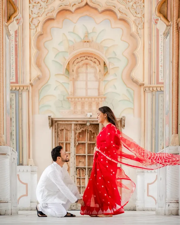 A truly royal pose at Jaipur's Patrika Gate. The groom kneels before his bride, whose red saree flows beautifully against the intricate architecture, creating a moment of pure grandeur.
