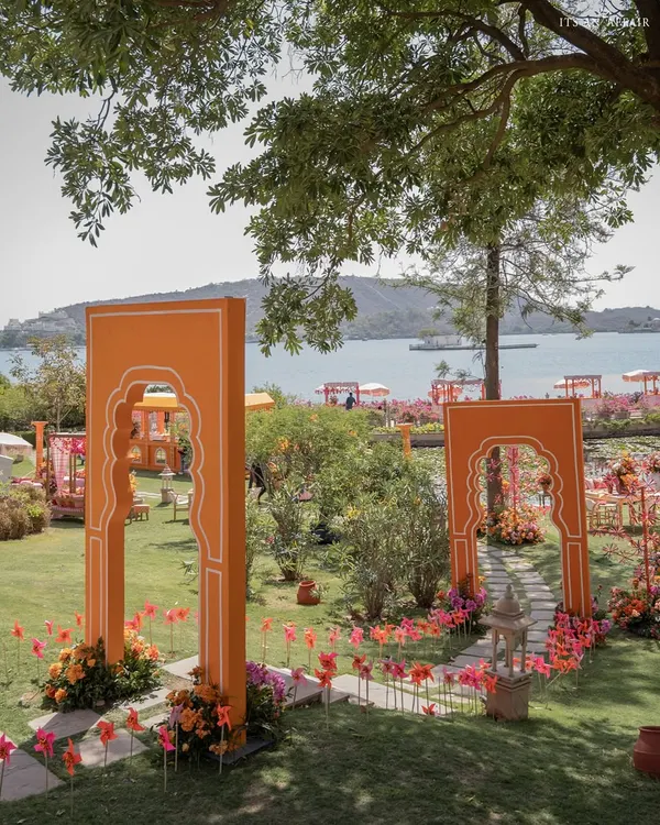 The entrance to the 'Pyaar ka Pind' Haldi, with vibrant orange arches framing a stunning view of Lake Pichola in Udaipur. This sets the stage for a colorful, destination celebration.