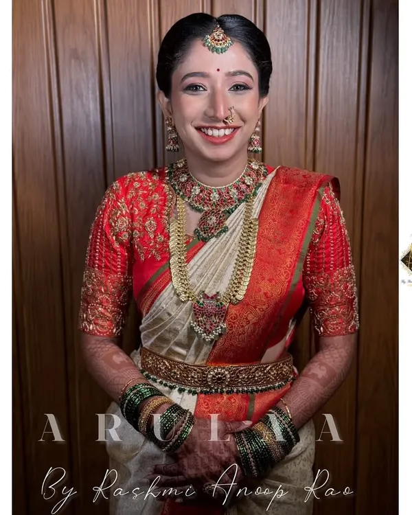 A radiant bride in her muhurtham attire. This red blouse features dense, all-over zardosi embroidery, paired with a classic off-white Kanjivaram saree for a timeless South Indian bridal look.