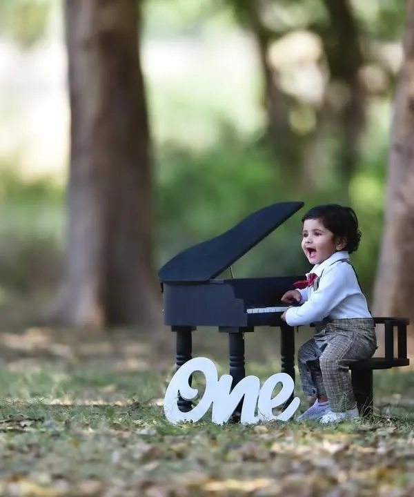 A joyful first birthday photoshoot in a park, featuring the birthday boy having fun with a miniature piano prop. The "One" sign adds a perfect touch to celebrate the milestone.