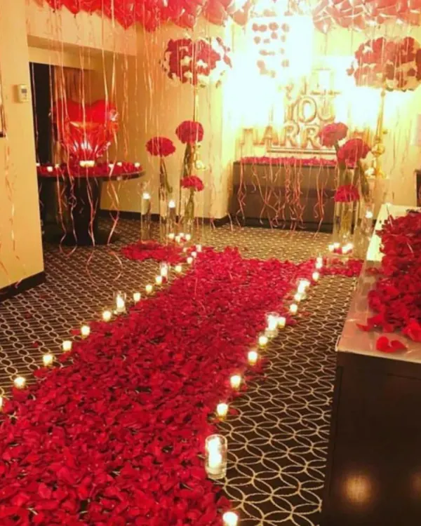 A luxurious room decoration for a proposal, featuring a dense pathway of red rose petals, candlelights, and red foil balloons creating a deeply romantic ambiance.