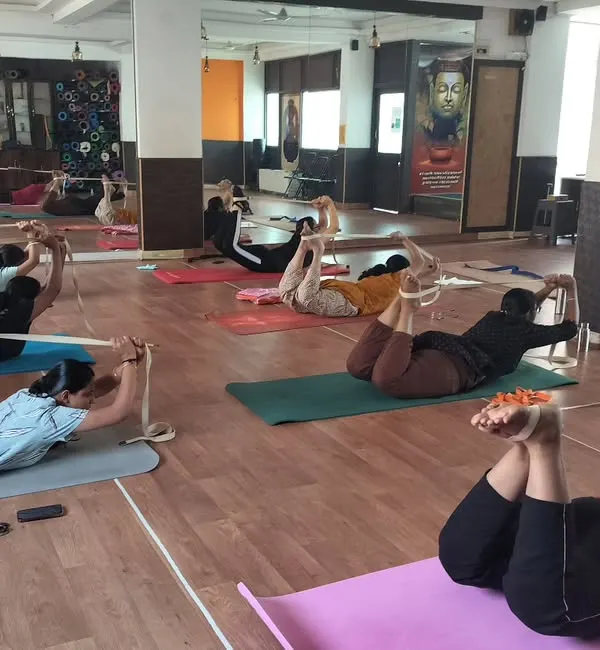 In this class, students are using yoga straps to assist with Dhanurasana (Bow Pose). The strap helps to get a better lift and a deeper opening in the chest and shoulders, making the pose more accessible.