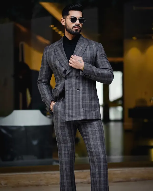 A sharp, full-length portrait from a men's formal wear photoshoot. The model is styled in a grey plaid double-breasted suit and black turtleneck, captured with a focus on the suit's tailoring and fit.