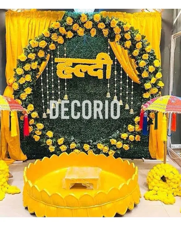 A complete Haldi ceremony stage with a lush green panel and a bright yellow floral arch. The setup includes traditional Rajasthani umbrellas and a central low seating area for the main rituals.