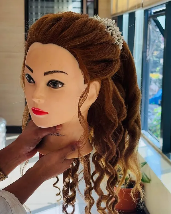 A mannequin head is used to demonstrate a romantic half-up, half-down hairstyle. This features soft curls and a twisted crown, adorned with delicate white flowers, perfect for a special occasion.