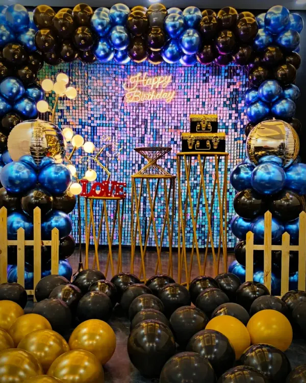 A bold and stylish birthday setup featuring a full frame of blue, black, and gold chrome balloons. The design is enhanced by a shimmering backdrop, a "Happy Birthday" neon sign, and gold disco ball accents for that extra sparkle.
