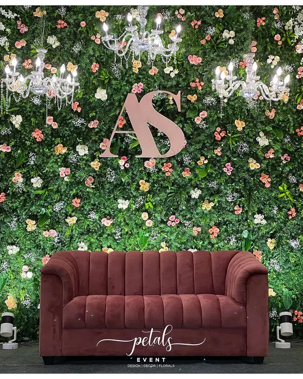 This reception stage is a perfect blend of nature and glamour. A lush green fern wall is adorned with pastel flowers, sparkling chandeliers, and the couple's initials, with a luxurious velvet sofa taking center stage.