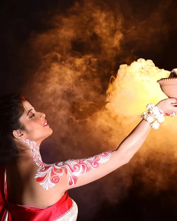 A creative shot for Durga Puja, where the model holds a dhunuchi (incense burner). The traditional Alpona body art on her arm is prominently displayed, glowing against the smoky background.