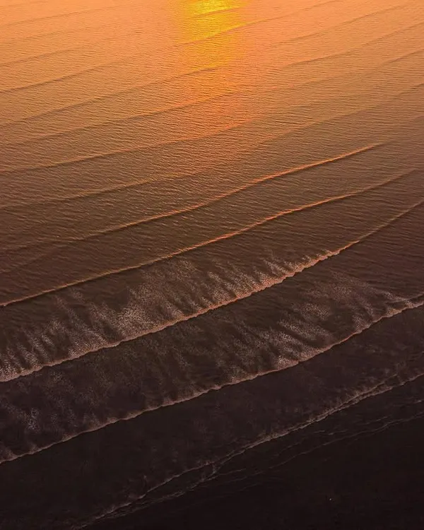 A close-up aerial shot of the ocean at sunset, where the low sun creates a golden reflection and highlights the texture of the parallel waves.