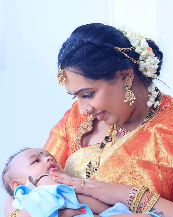 A precious moment between mother and child at her baby boy's naming ceremony. Her makeup is soft and radiant, with a beautiful floral bun, perfect for this heartfelt occasion.