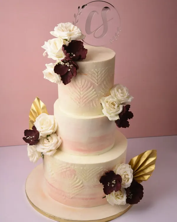 A stunning three-tier wedding cake with a soft peach ombré effect and delicate leaf-stamped texture. It's adorned with white and deep burgundy sugar flowers and gold leaf accents for a touch of modern romance.