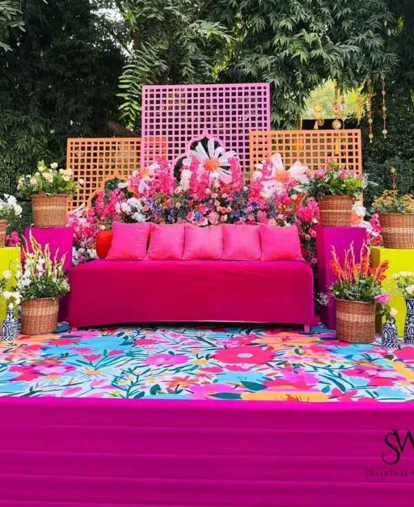 A vibrant and colorful Mehendi or Haldi seating area. The bright pink sofa, floral printed flooring, and abundant flowers in wicker baskets create a festive and cheerful atmosphere.