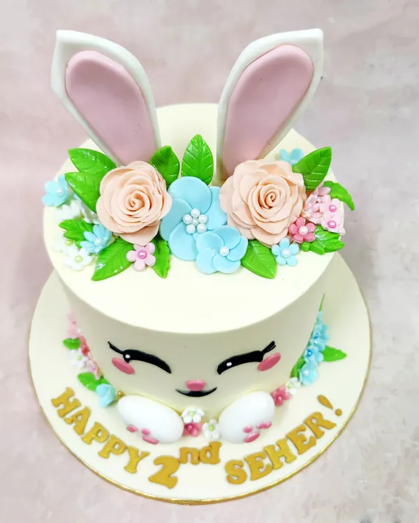 We craft pure pastel joy with this adorable bunny face cake. Perfect for a second birthday, this design features a sweet bunny face with large ears peeking out from a crown of delicate, handcrafted sugar flowers in soft peach and blue.