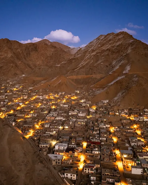 An aerial view of a town in Ladakh as dusk settles, with lights beginning to glow in the valley. This shot captures the quiet beauty of human settlement nestled amongst the towering, barren mountains of the Himalayas.