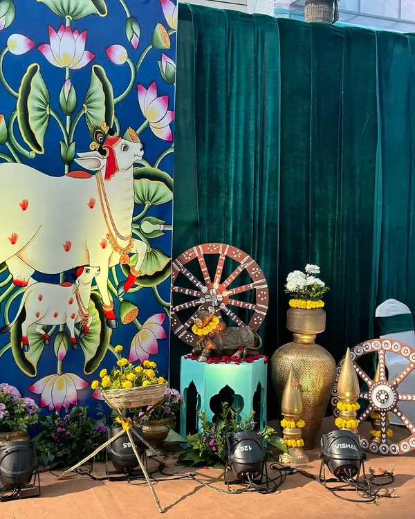 A beautiful setup for a Radha Krishna themed event, featuring a custom backdrop with Pichwai art of a cow and calf. The decor is complemented by brass pots, painted wheels, and marigold flowers.