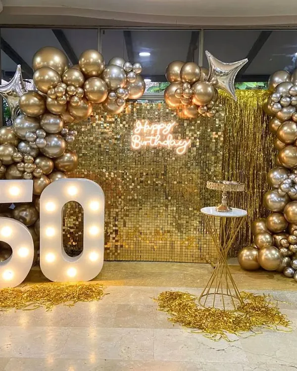 A glamorous 50th birthday setup featuring a gold sequin wall that catches the light beautifully. The scene is framed by a sophisticated balloon arch in metallic gold and silver, with large, illuminated marquee numbers proudly displaying the age.