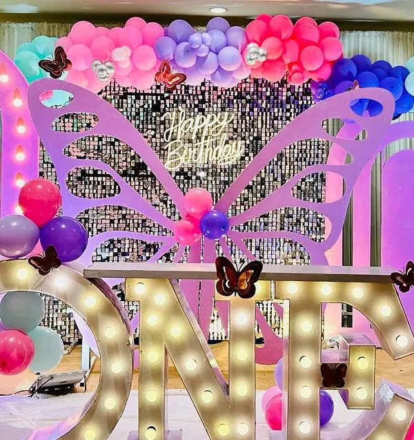 A magical first birthday setup combining a butterfly theme with large "ONE" marquee letters. The decor features a silver shimmer wall, colorful balloon arrangements in teal, pink, and purple, and detailed butterfly cutouts.