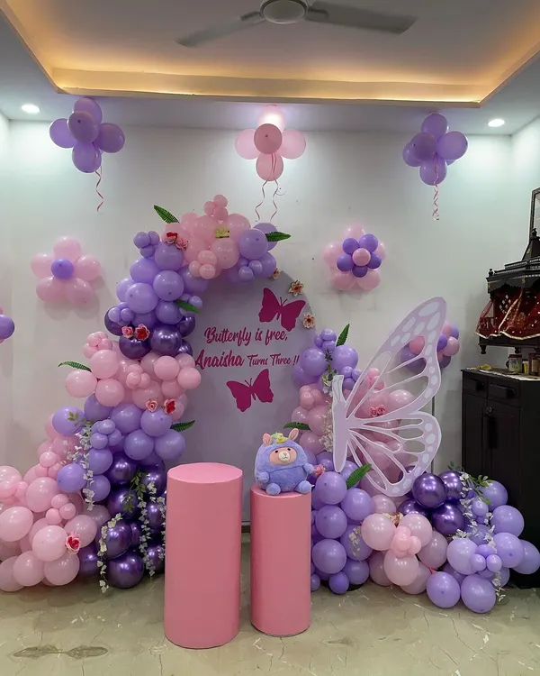 A beautiful butterfly-themed party in shades of purple and pink. This setup features a balloon arch around a custom backdrop, large butterfly wings for photos, and flower-shaped balloon clusters on the wall.