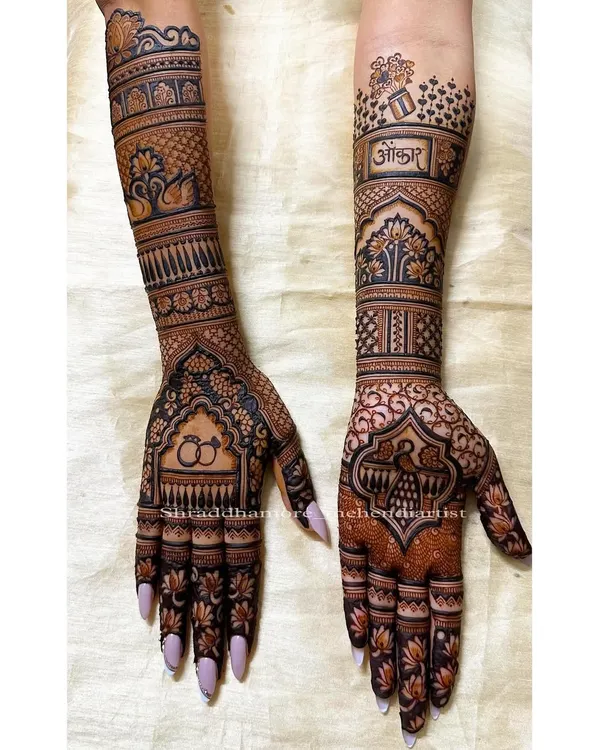 This full-arm bridal mehendi was created for Ankita, featuring her partner's name 'Omkar' on one arm and engagement rings on the other. The design combines traditional elements like swans and peacocks with personal touches.