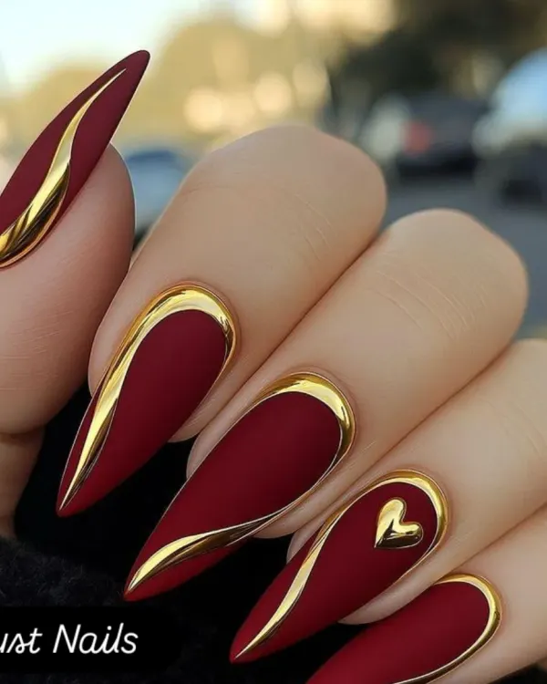 A dramatic and luxurious look featuring long, stiletto-shaped extensions with a matte crimson base and bold, raised gold chrome accents. One nail features a delicate gold heart, adding a touch of romance to this powerful design.