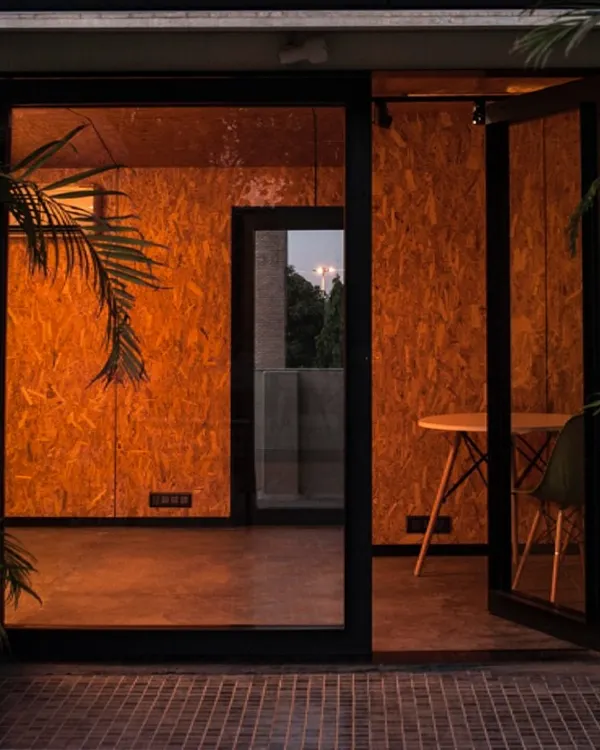 The entrance to a small workspace at dusk. The warm interior light glows through the full-height glass door, creating an inviting threshold between inside and out.