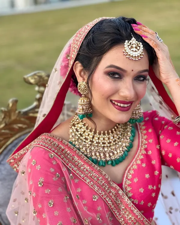 A picture-perfect bride with a dazzling smile. Her makeup features smoky eyes and a bold berry lip, beautifully contrasting with her bright pink lehenga and emerald green jewellery.