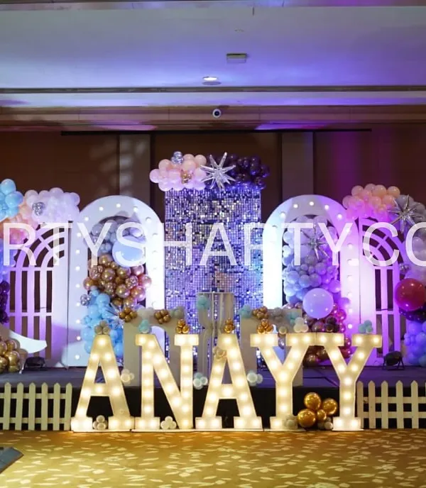 This adorable teddy bear stage is perfect for a first birthday. The setup features two giant teddy bear props, a sparkling sequin backdrop, and a white picket fence in front of the illuminated name 'ANAYY', creating a cute and cuddly atmosphere.