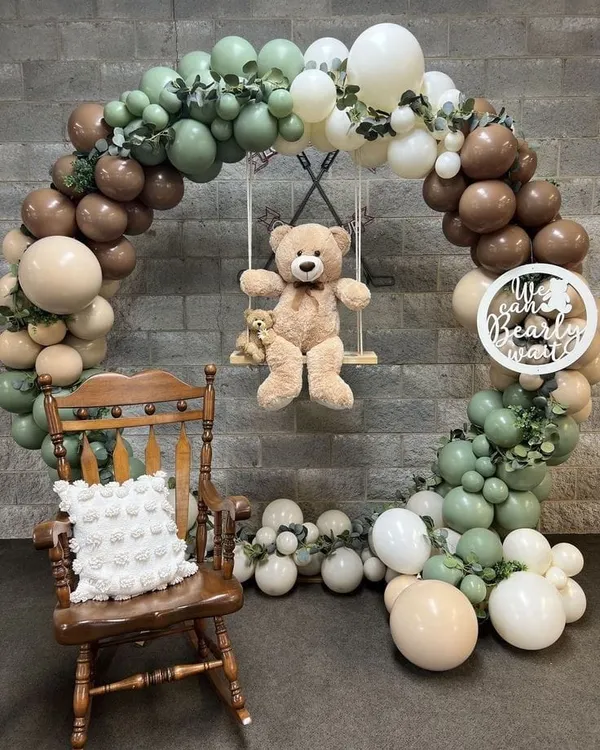 A rustic teddy bear theme with a teddy on a swing inside a balloon arch. The arch uses sage green, beige, and brown balloons, perfect for a "We Can Bearly Wait" baby shower.
