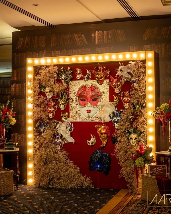 The centerpiece of our speakeasy masquerade photo booth. A large, ornate red mask is surrounded by a collection of Venetian-style masks, framed by vintage marquee lights to create a dramatic and theatrical entrance for guests.