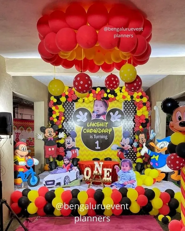 This is a complete Mickey Mouse themed first birthday party I created. The setup includes a personalized backdrop with the child's photo, multiple character cutouts, and coordinated red, yellow, and black balloon decorations for a fun and immersive experience.