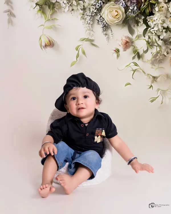 A simple and elegant studio portrait. The baby is dressed in a stylish dark shirt and cap, set against a clean white background with a delicate floral arrangement overhead.