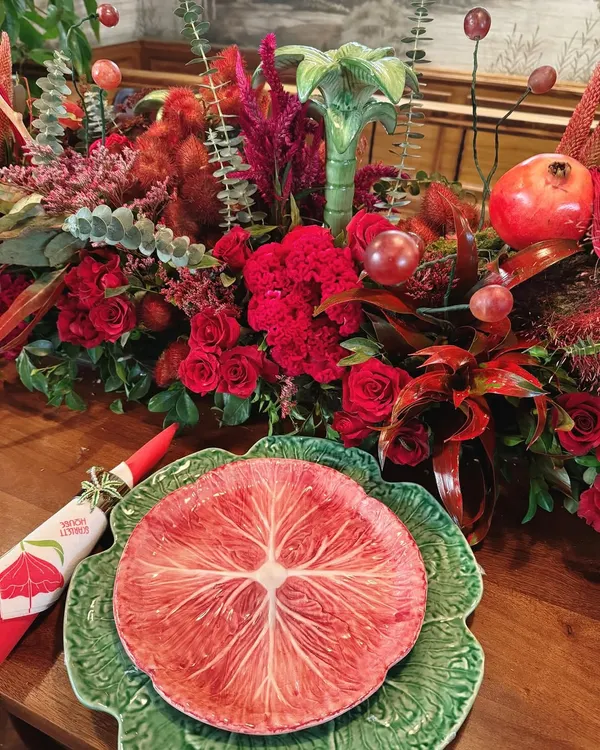A close-up of a place setting from our "Scarlet House" theme. A vibrant, custom-printed plate sits atop a green charger, complemented by a bold red napkin and a backdrop of lush, textured red florals.