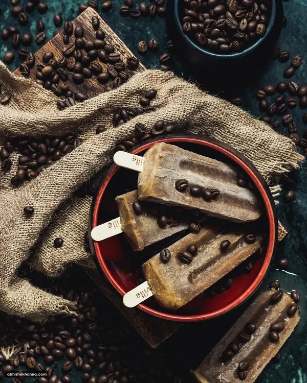 Coffee-flavored popsicles styled in a dark, moody setting with scattered coffee beans. This conceptual shot is designed to be artistic and attention-grabbing.