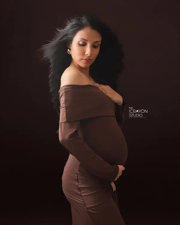 A fine art portrait against a rich, dark backdrop. The soft light highlights the curve of her bump and the gentle movement of her hair, creating a poetic and graceful image.