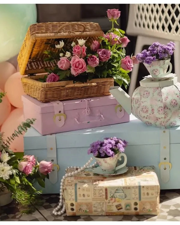 A beautiful collection of props for a vintage tea party theme. The display includes pastel-colored storage trunks, a wicker picnic basket filled with roses, and a floral teapot centerpiece.