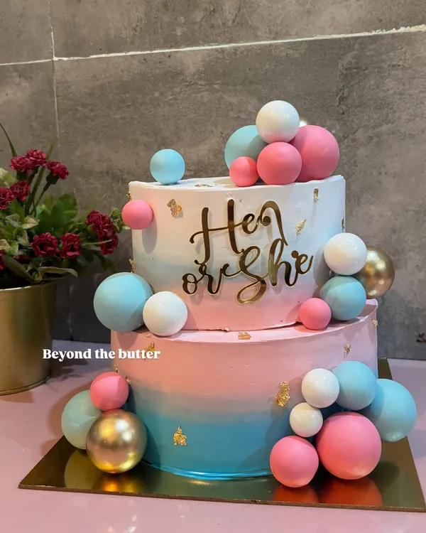 He or She? This two tier gender reveal cake is full of suspense and excitement, decorated with pink, blue, and gold chocolate spheres. It's a fun way to share your big news with family and friends.