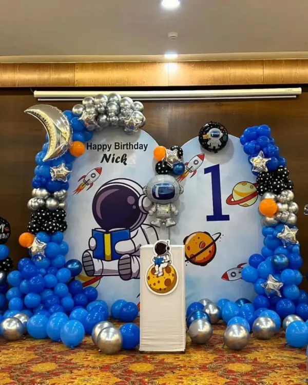 A complete astronaut-themed stage for Nick's first birthday. This setup includes a large custom backdrop with space graphics, a full balloon garland with silver and orange accents, and a themed cake stand.