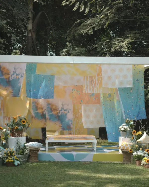A full view of the unique "Dhobi Ghat" themed Haldi setup. The backdrop of colorful tie-dyed fabrics, combined with a patterned stage floor and sunflower arrangements, creates a vibrant and artistic atmosphere.