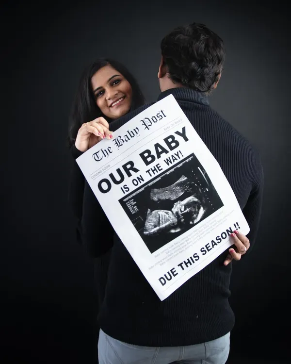 A playful and creative announcement. The mom-to-be peeks over her partner's shoulder while he holds a custom newspaper prop announcing their baby is on the way.