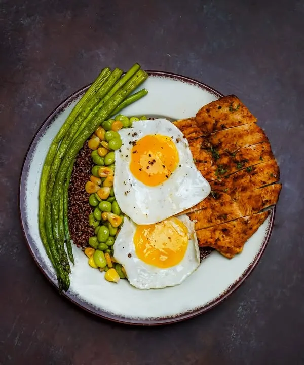 A high-protein meal with grilled chicken, two sunny-side-up eggs, quinoa, and fresh green beans, shot with dramatic lighting.