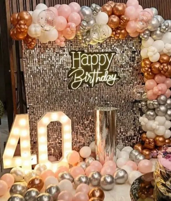 A glamorous 40th birthday setup with a rose gold, pink, and silver balloon arch over a shimmering sequin wall, complete with a neon sign and large marquee numbers.