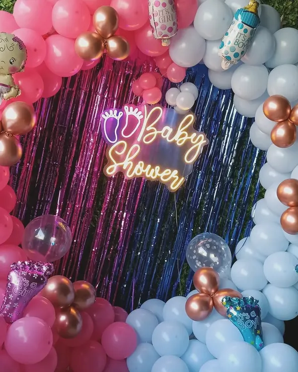 This is a popular baby shower decoration. It features a full ring of pink and blue balloons, a shiny backdrop, a "Baby Shower" neon sign, and cute baby-themed foil balloons.