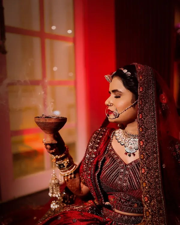 A serene bridal portrait showing the bride in her red lehenga, holding a traditional bowl with incense smoke. The warm, red lighting creates a spiritual and deeply cultural atmosphere for this wedding day photo.
