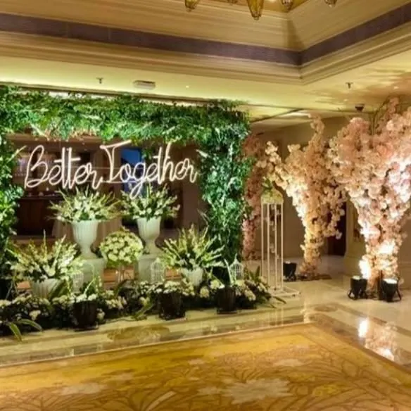 A beautiful photo corner for an engagement party with a 'Better Together' neon sign. The backdrop of lush greenery and cherry blossom trees creates a romantic and whimsical setting.