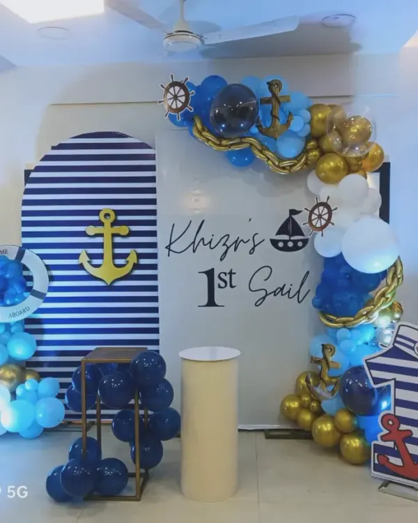 A nautical-themed decoration for "Khiza's 1st Sail". This setup features a striped backdrop, an anchor cutout, a ship's wheel, and a blue and gold balloon arch, perfect for a little sailor's party.
