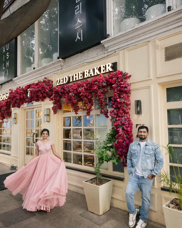 A modern fairytale scene in front of a beautiful bakery. The contrast between her flowing pink gown and his casual denim creates a stylish, contemporary pre-wedding portrait.