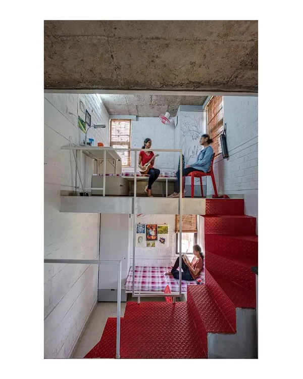 The interior of a room in our 'Stacked Student Housing' project, showing how a split-level design maximizes space. The lofted study area and lower sleeping zone create distinct functional areas within a compact footprint, connected by a bold red steel staircase.
