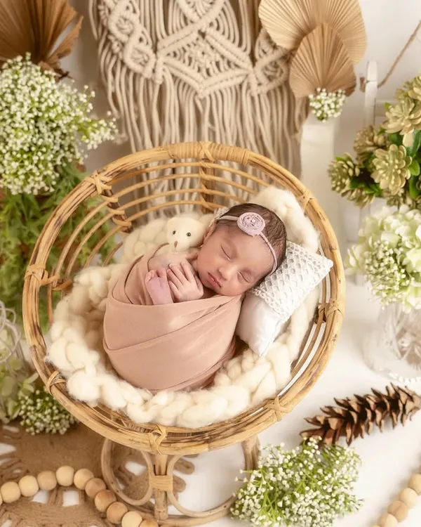 This precious baby is swaddled and sleeping in a stylish rattan chair. The background features a macrame wall hanging, dried leaves, and pinecones, creating a complete boho chic look.