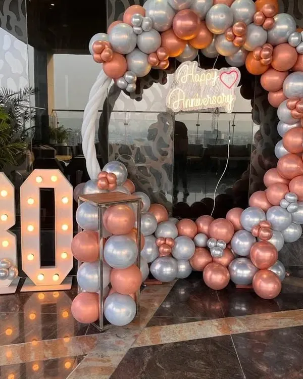 Celebrating a 30th anniversary with a stunning balloon arch against a city skyline. This setup includes a large circular arch in rose gold and silver chrome balloons, a custom 'Happy Anniversary' neon sign, and large light-up marquee numbers.